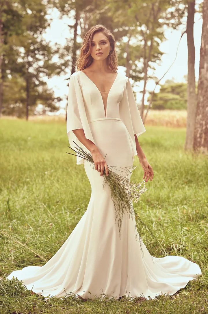 Woman in a white wedding dress standing in a grassy field with trees in the background
