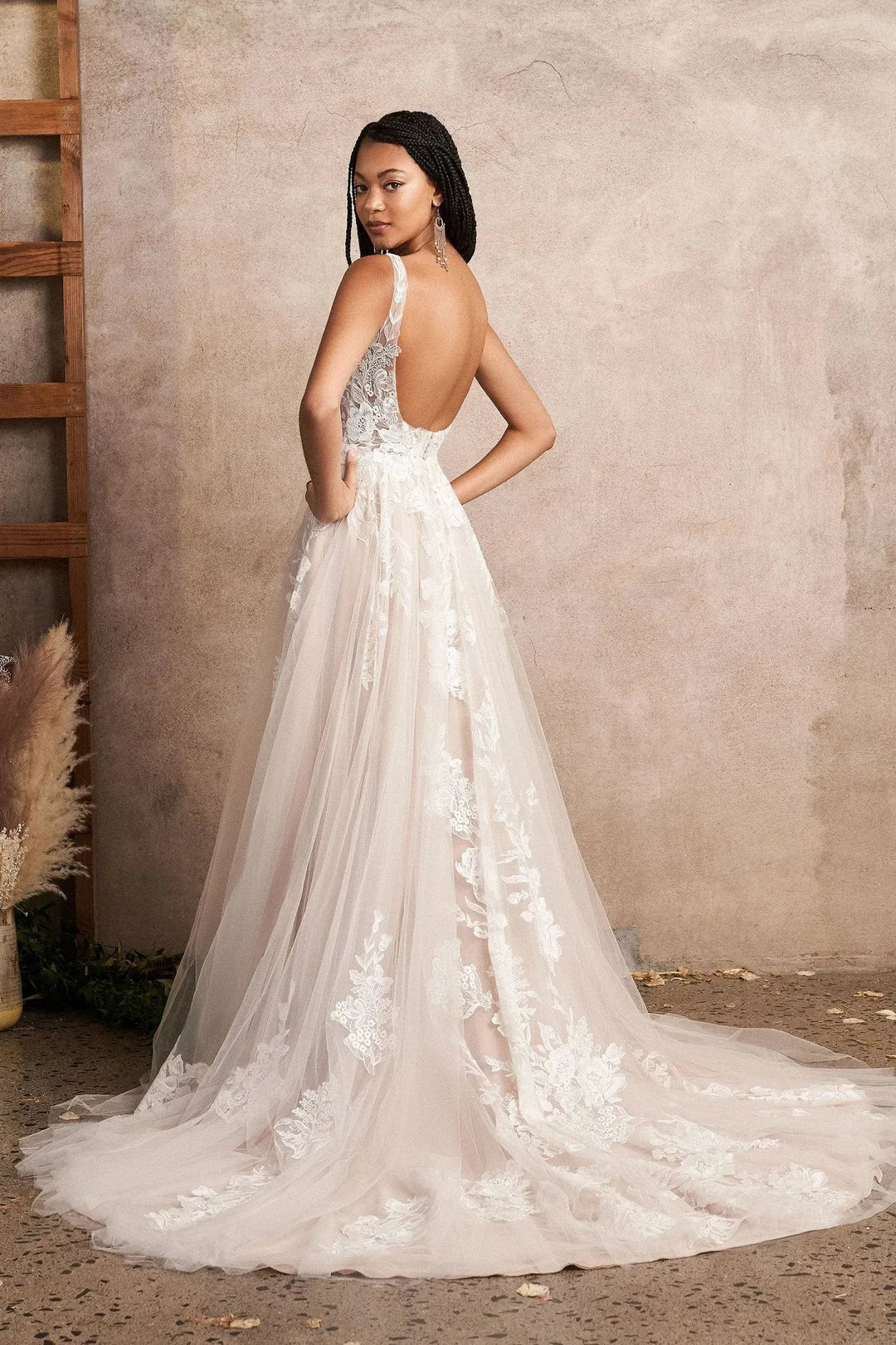 Woman wearing a white lace wedding dress with an open back against a neutral wall.