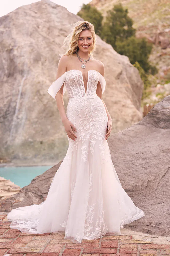 A woman wearing a strapless off-the-shoulder mermaid-style wedding gown with lace and tulle detailing, standing outdoors on a stone path with large rock formations and turquoise water in the background.