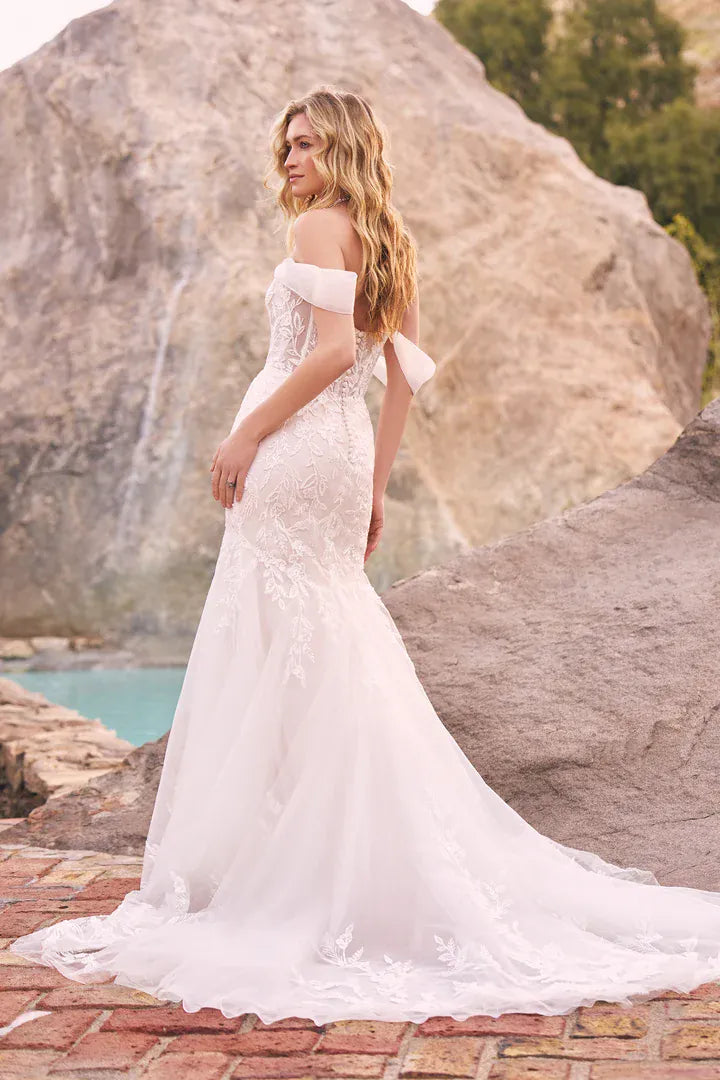 A woman wearing a white off-the-shoulder lace mermaid wedding gown stands outdoors on a stone patio with large rock formations and turquoise water in the background.