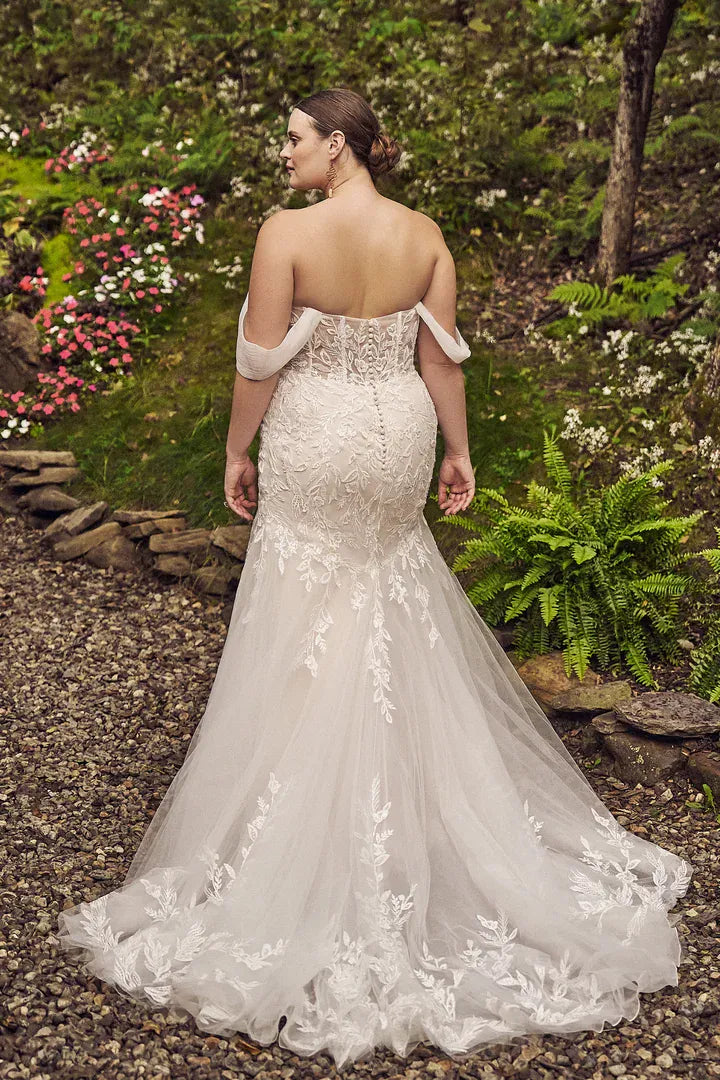 A woman wearing a white off-the-shoulder lace wedding gown is photographed from the back in an outdoor garden setting with gravel ground and green plants.