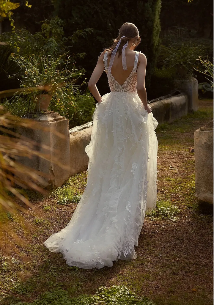 Woman in a white wedding dress standing in a garden setting with trees and stone walls.
