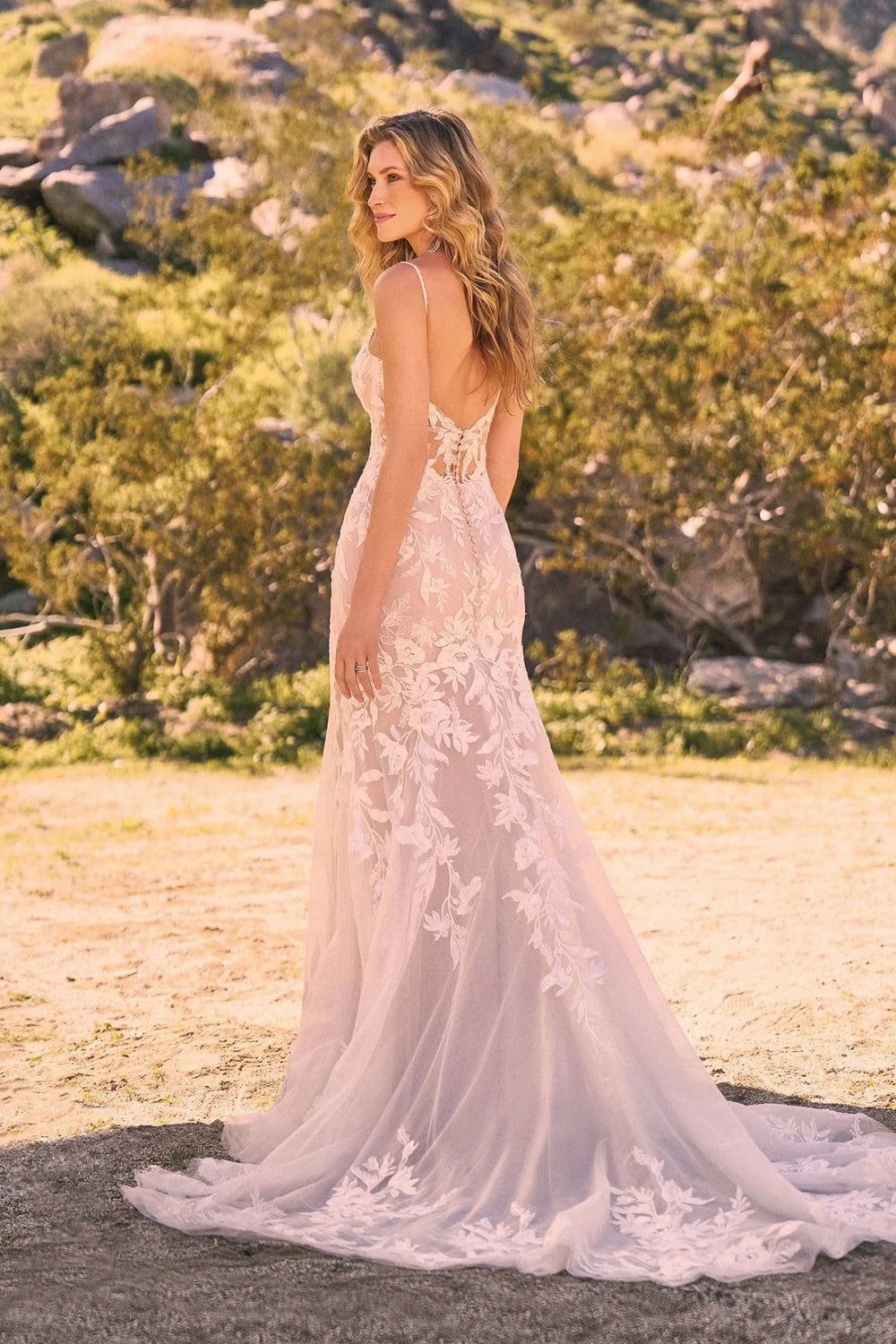 Woman in a white lace wedding dress standing in a natural setting with trees and rocks.