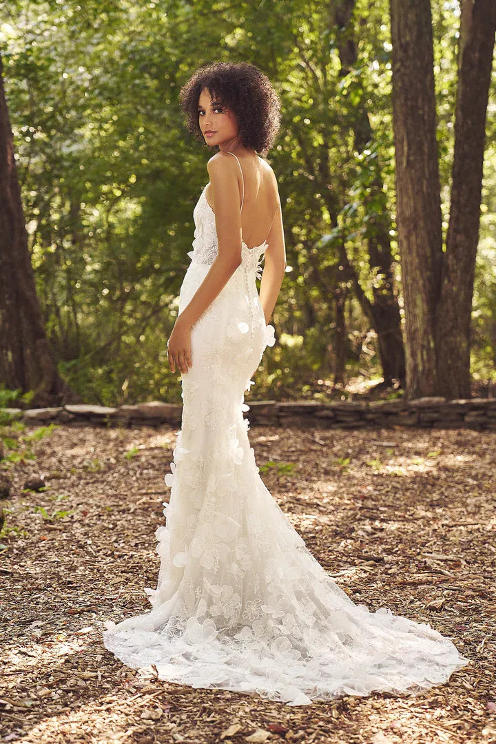 A woman wearing a white lace wedding gown stands in a wooded outdoor setting, photographed from the back as she looks over her shoulder. The dress has a fitted silhouette with floral lace appliqué and a long train on a leaf-covered forest floor.