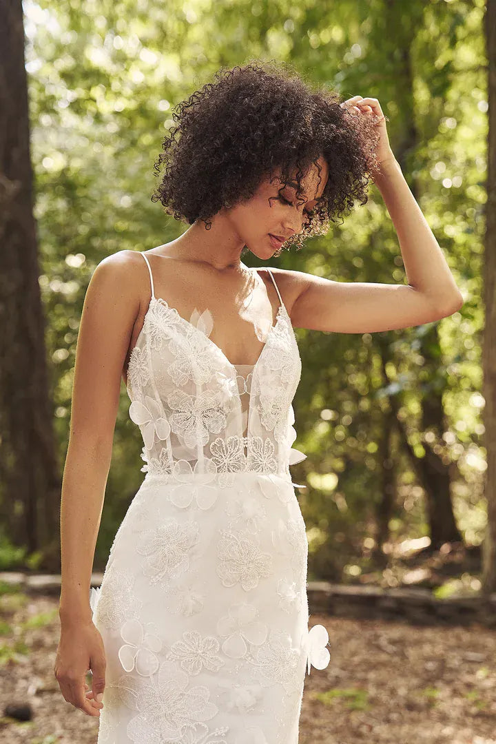 A woman wearing a white lace spaghetti-strap dress stands outdoors in a sunlit wooded setting, with her hand touching her hair.