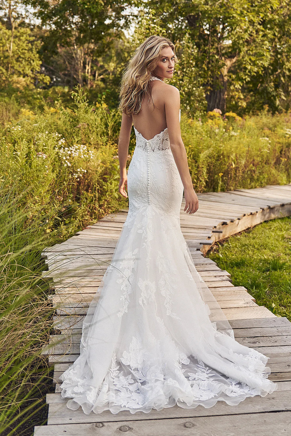 Woman in a white lace wedding dress standing on a wooden path in a natural setting.