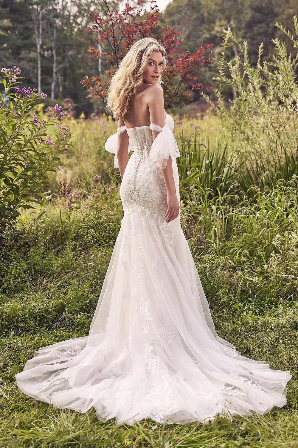 Woman in a white wedding dress standing in a natural setting with greenery and flowers.