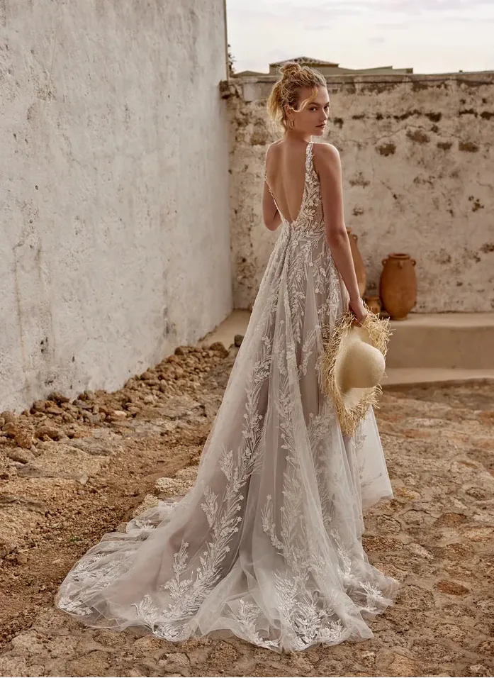 A woman wearing a sleeveless lace wedding gown with a deep V back and a long train, standing outdoors against rustic stone walls and holding a straw hat.