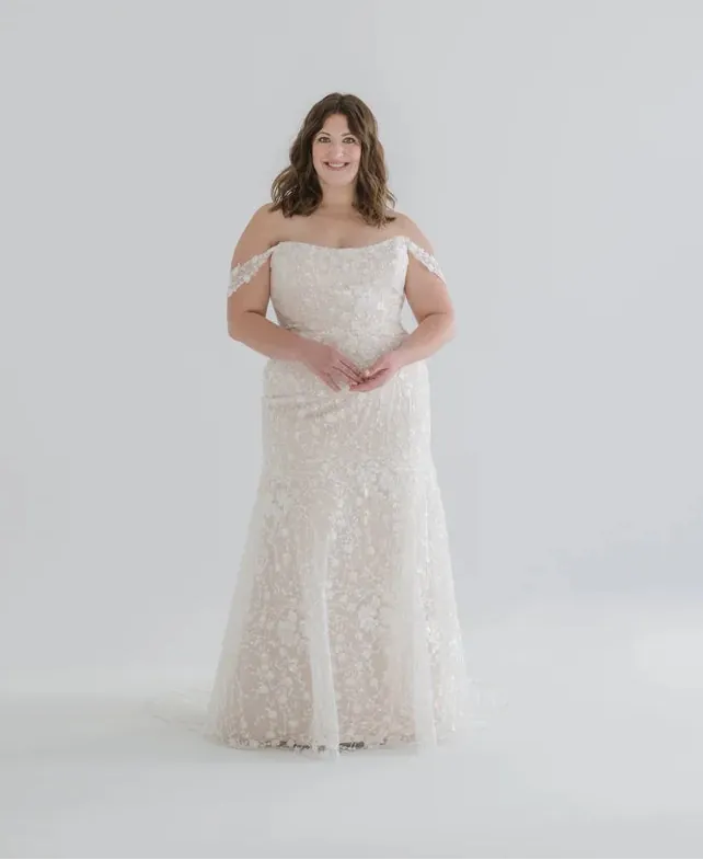 A woman modeling an off-the-shoulder lace wedding gown in a cream or ivory shade, standing against a seamless light gray studio background.