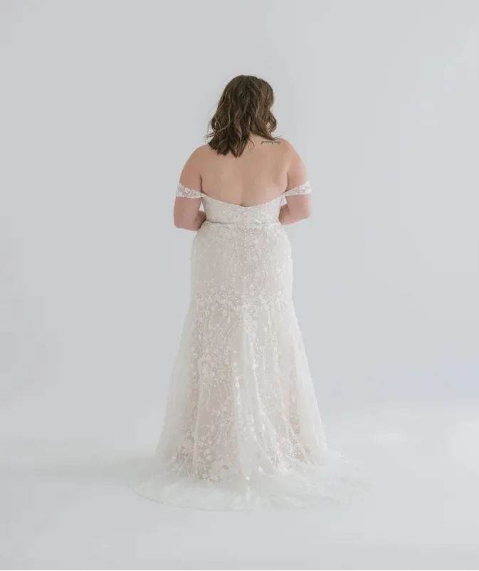 A woman viewed from behind wearing an off-the-shoulder lace wedding gown with a fitted bodice and full tulle skirt, photographed against a seamless light gray studio background.