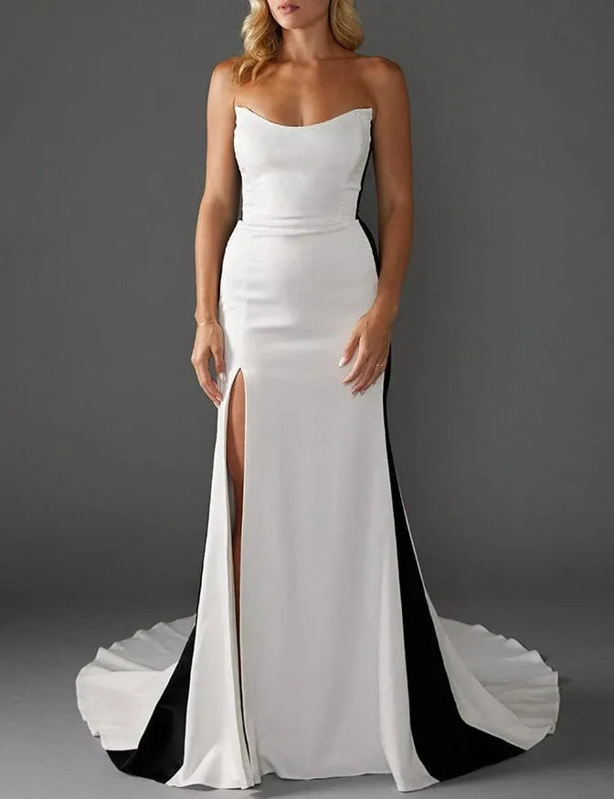 A woman models a strapless white floor-length gown with a sweetheart neckline, fitted silhouette, thigh-high slit, and a dramatic black-and-white draped train against a dark gray studio background.