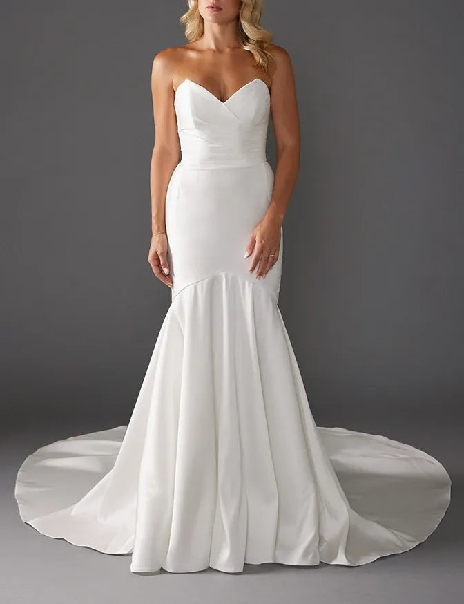 A woman modeling a strapless white mermaid-style wedding gown with a sweetheart neckline, fitted bodice, flared skirt, and long train against a dark gray studio background.