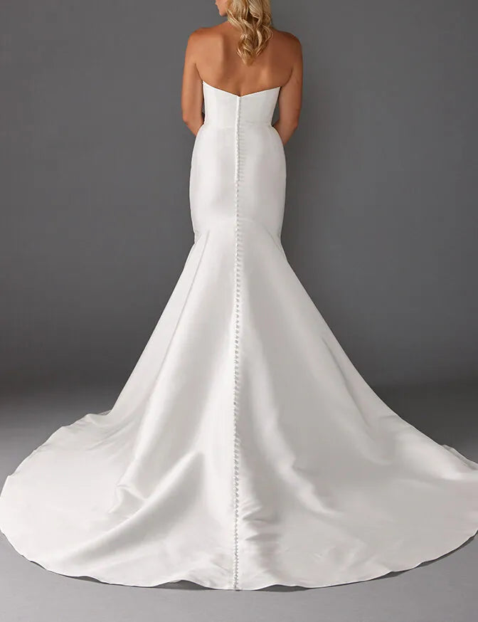 A strapless white mermaid-style wedding gown shown from the back, featuring a structured back with a central vertical seam and a full satin train, photographed against a dark gray studio background.