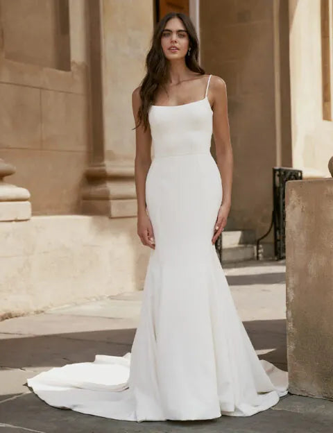 A woman modeling a sleeveless white mermaid-style wedding gown with thin straps, a fitted bodice, and a slight train, standing outdoors in front of a beige stone building.