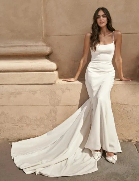 A woman wearing a sleeveless white mermaid-style wedding gown with thin straps, a fitted bodice, flared hem, and a long train, posed against a beige stone wall.