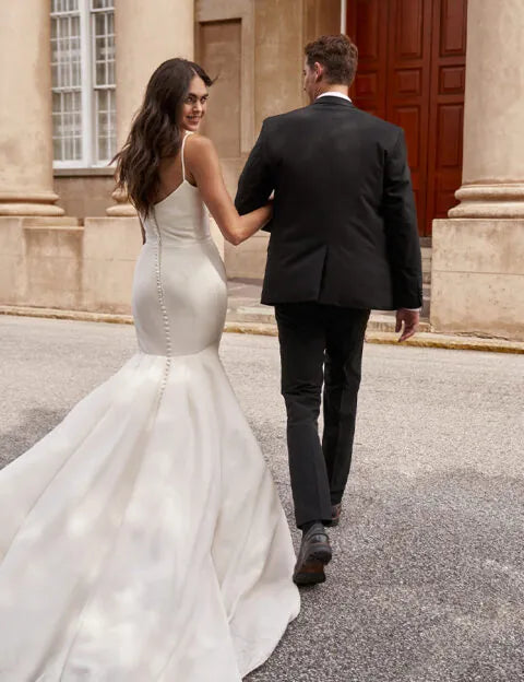 A bride in a fitted white mermaid-style wedding gown walks arm in arm with a groom in a black suit outdoors in front of a stone building with a large wooden door.