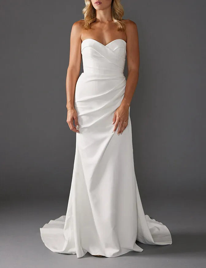 A woman modeling a strapless white wedding gown with a sweetheart neckline, ruched bodice, and a slight train against a dark gray studio background.