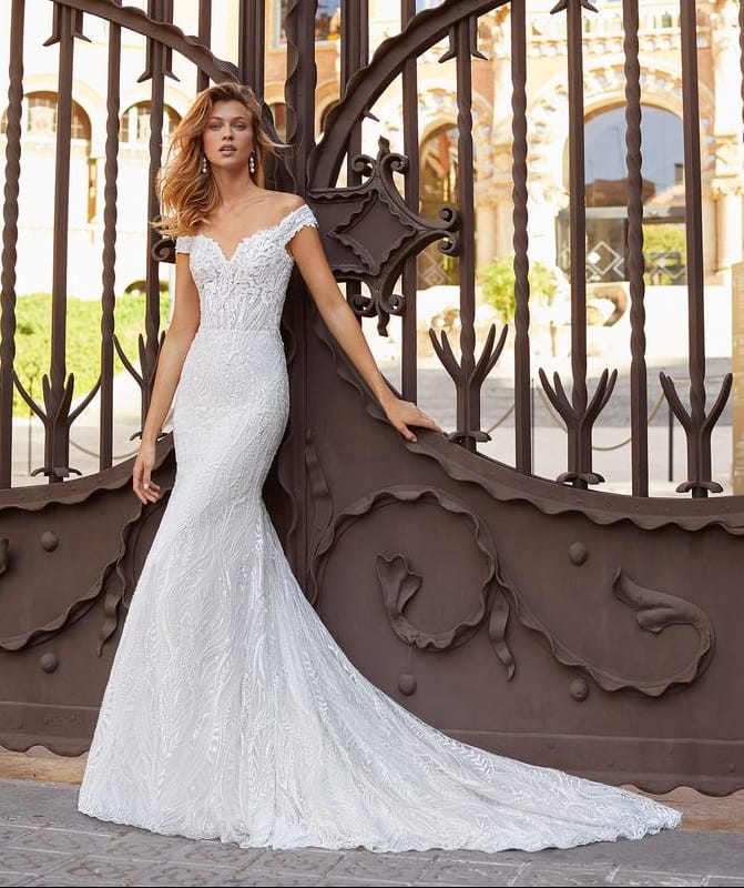 A woman wearing a white lace mermaid-style wedding gown stands in front of an ornate dark metal gate, with a building visible in the background.