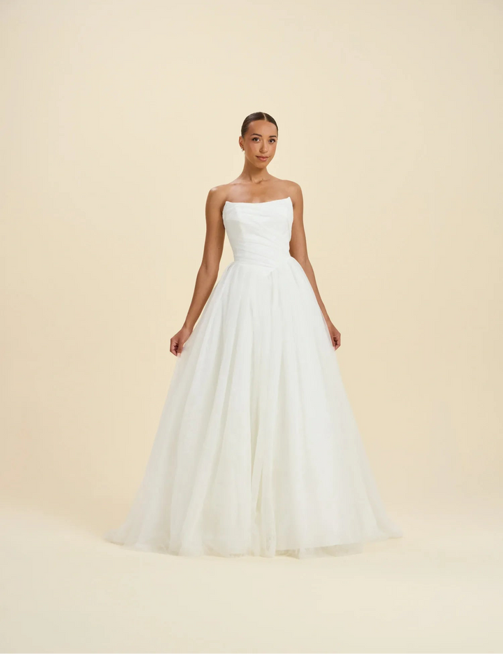 A woman modeling a strapless white wedding gown with a fitted bodice and a full, flowing tulle skirt against a plain warm beige studio background.