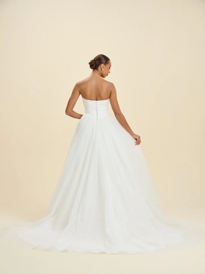 A woman modeling a strapless white wedding gown from the back, showing a structured bodice and a full, flowing tulle skirt against a warm cream studio background.