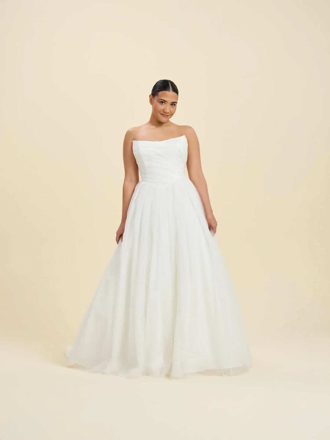 A woman modeling a strapless white wedding gown with a fitted bodice and a full, flowing tulle skirt against a plain beige studio background.