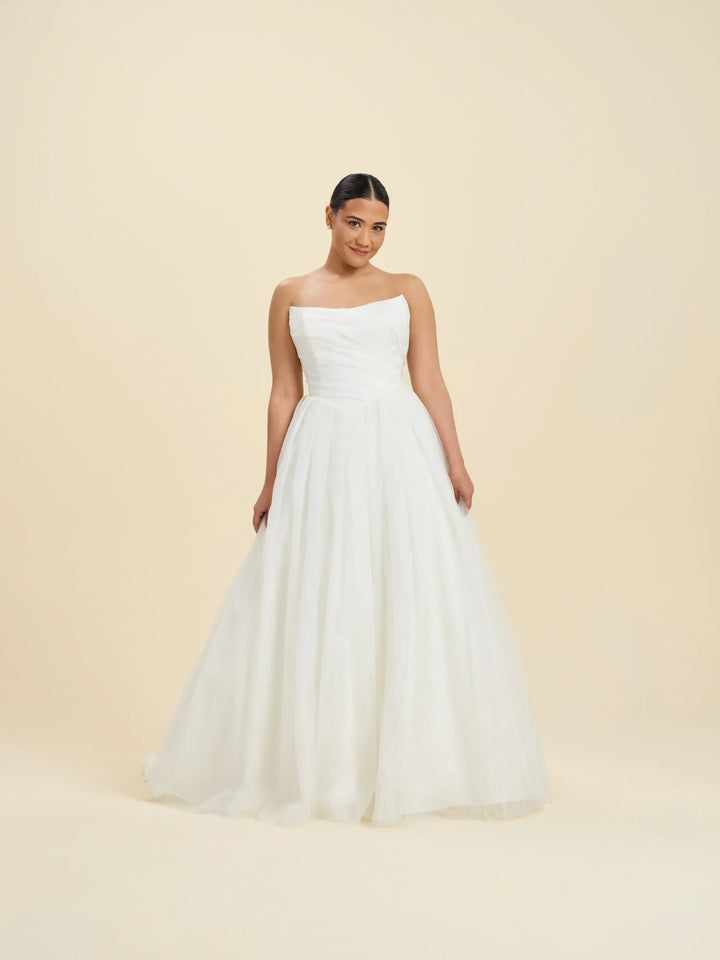 A woman modeling a strapless white wedding gown with a fitted bodice and a full, flowing tulle skirt against a plain beige studio background.