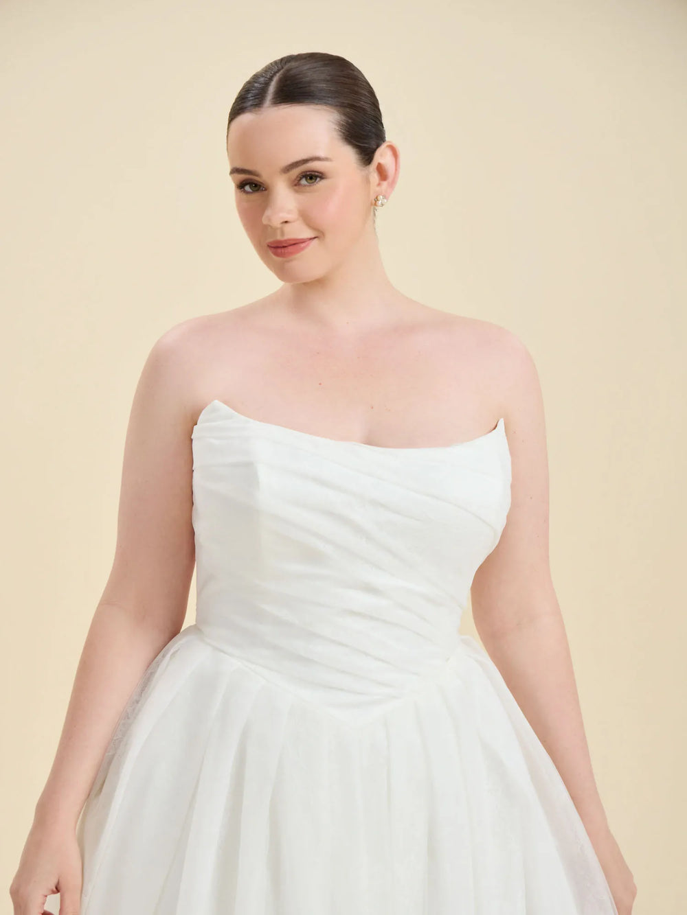 A woman modeling a strapless white wedding dress with a ruched bodice and full tulle skirt against a solid beige studio background.
