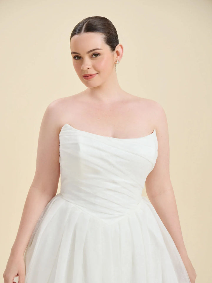 A woman modeling a strapless white wedding dress with a ruched bodice and full tulle skirt against a solid beige studio background.