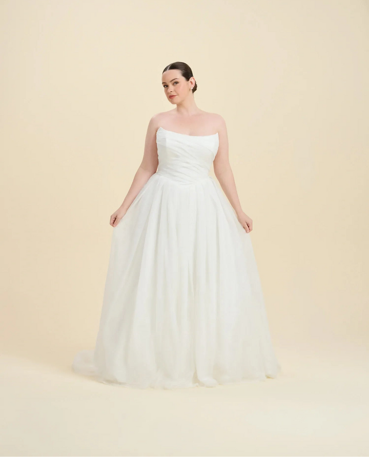A woman modeling a strapless white ball gown with a fitted bodice and full tulle skirt, standing against a plain warm beige studio background.