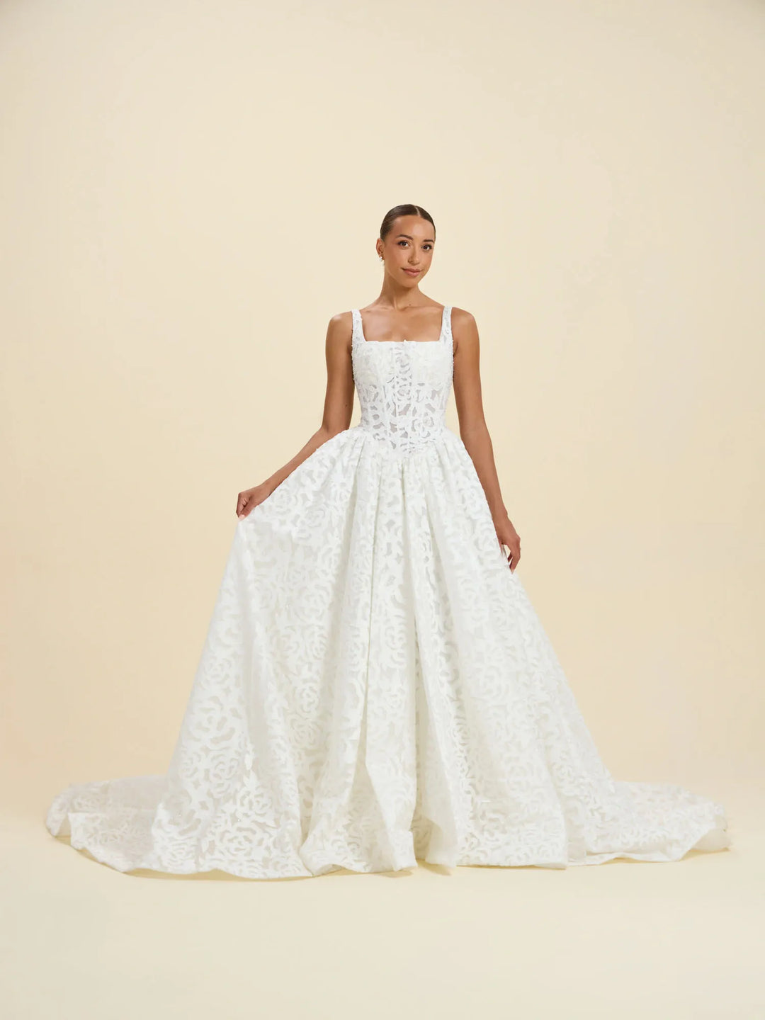A white lace wedding gown with a square neckline, thin straps, a fitted bodice, and a full flared skirt, shown on a model against a warm cream studio background.