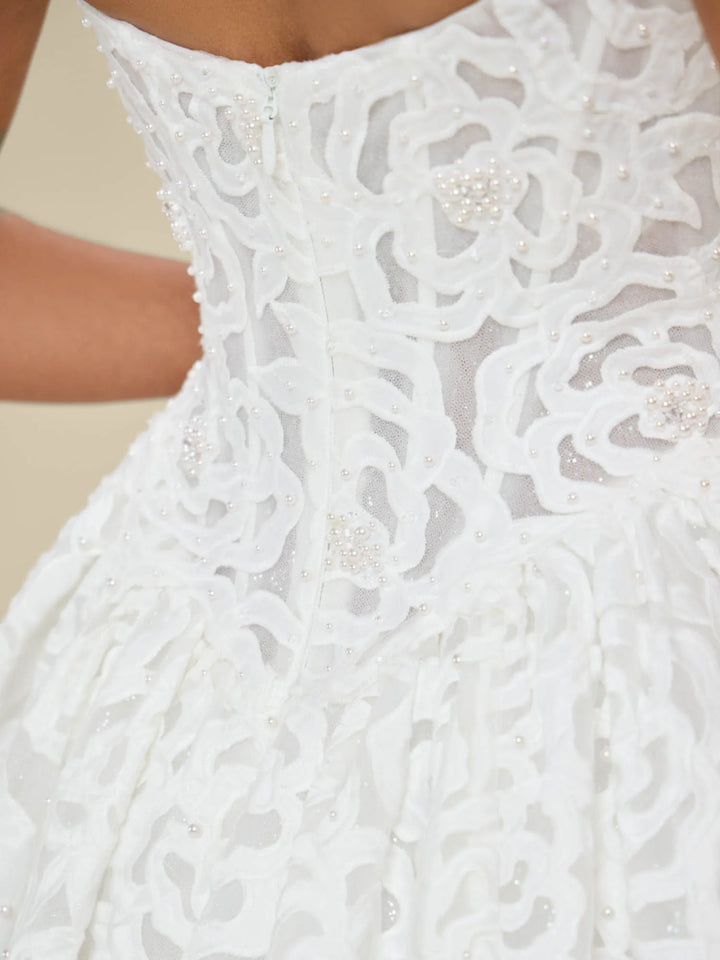 Close-up of a white lace bridal gown with floral embroidery, beaded details, and pearl-like embellishments on the bodice and skirt.