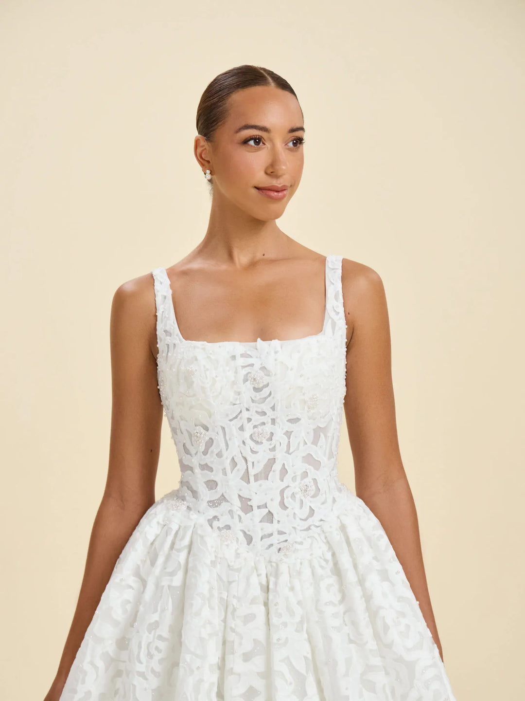 A woman modeling a white sleeveless lace dress with a square neckline, fitted bodice, and full skirt against a plain beige studio background.