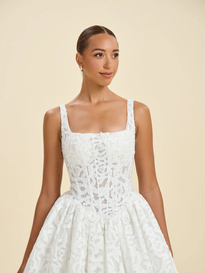 A woman modeling a white sleeveless lace dress with a square neckline, fitted bodice, and full skirt against a plain beige studio background.