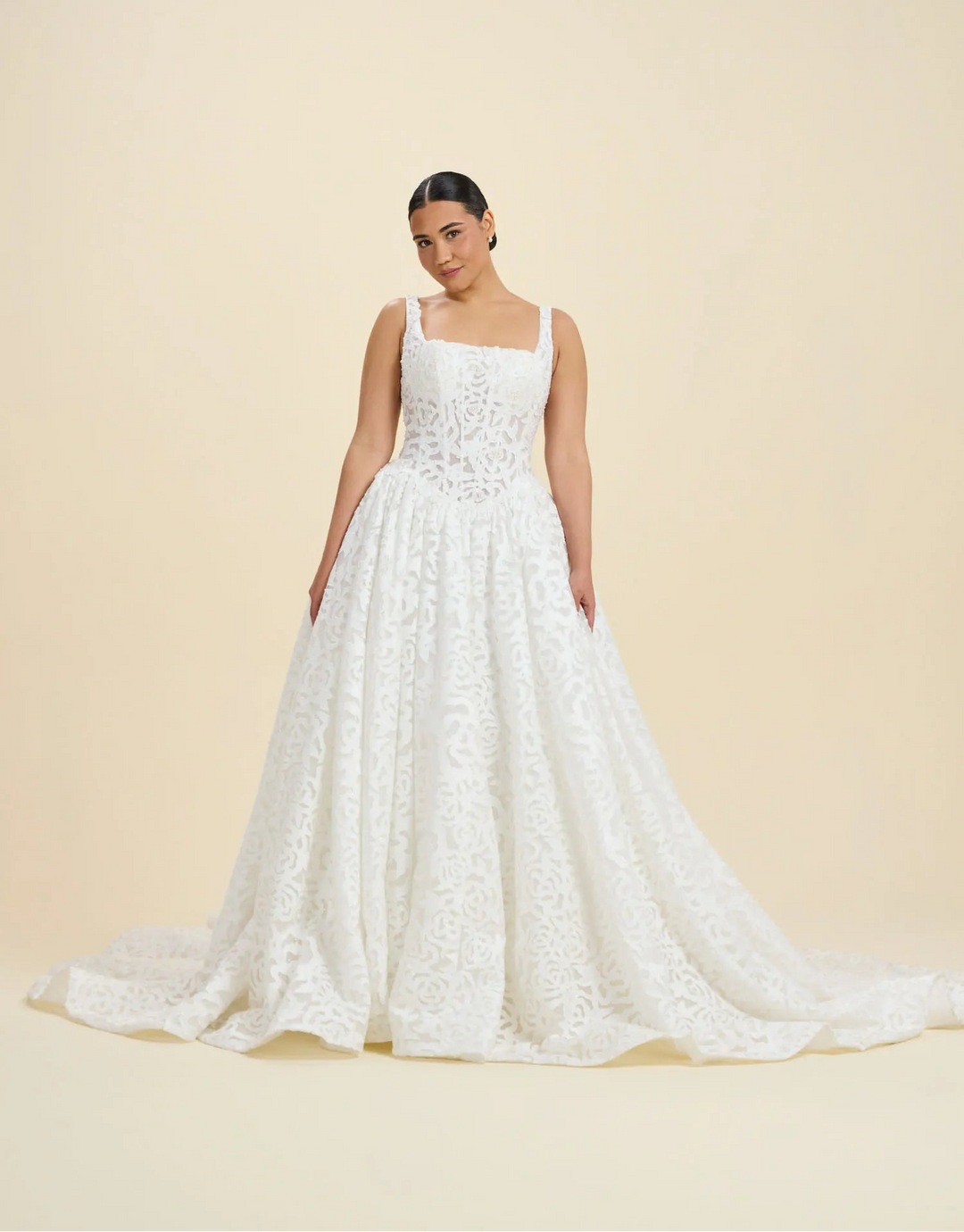 A woman modeling a white lace A-line wedding gown with thin straps, a square neckline, and a full skirt with a long train, set against a solid beige studio background.