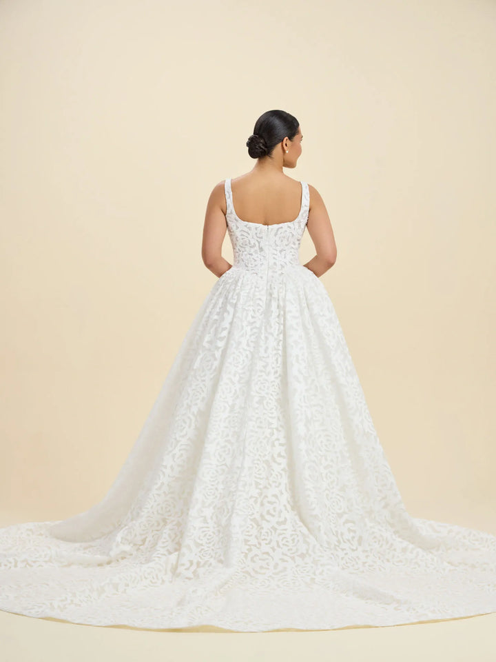 A woman viewed from the back wearing a white lace wedding gown with a full skirt and long train, standing against a plain warm beige studio background.