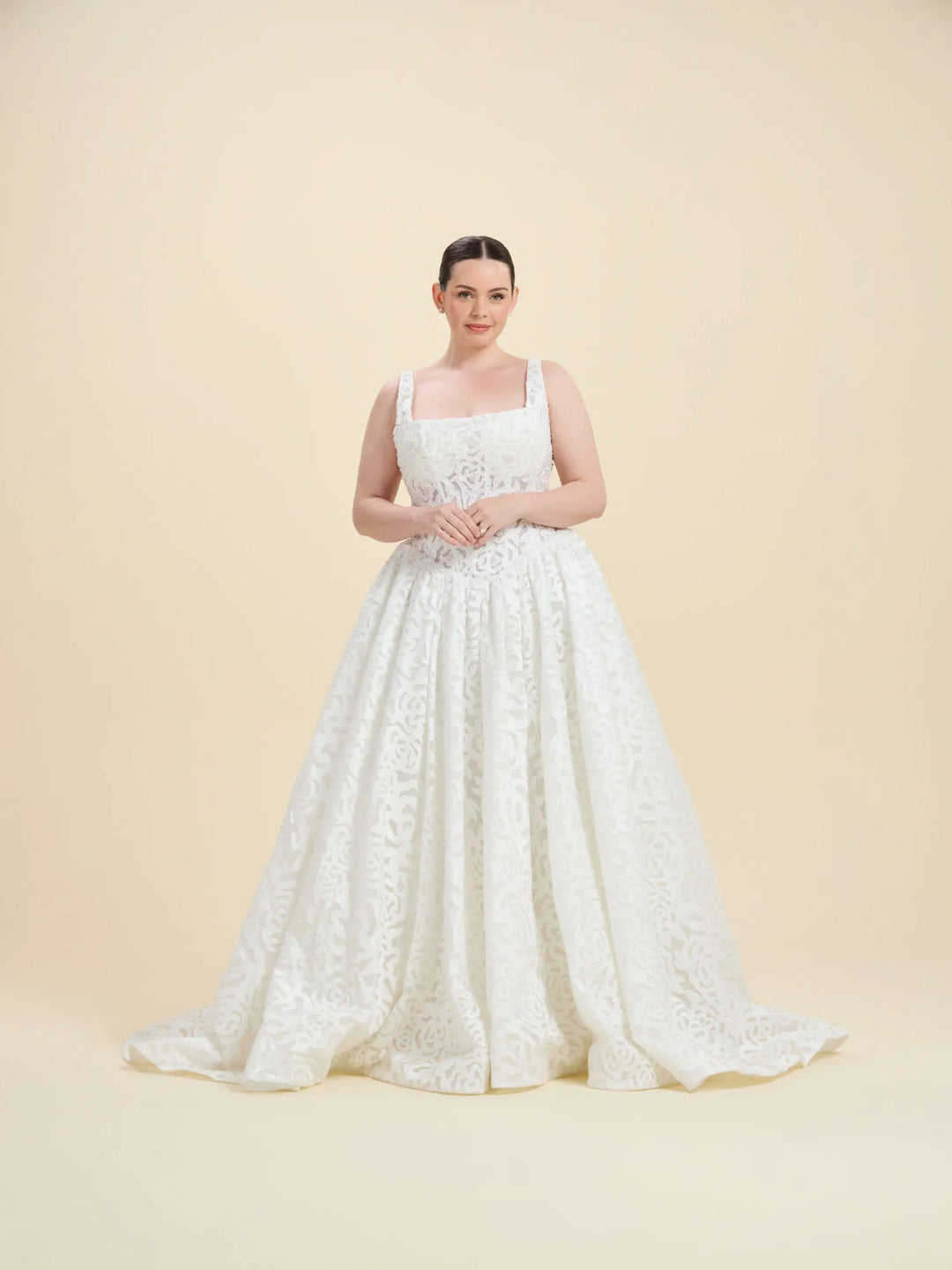 A woman modeling a white lace wedding gown with a square neckline, thin straps, and a full skirt, standing against a solid beige studio background.