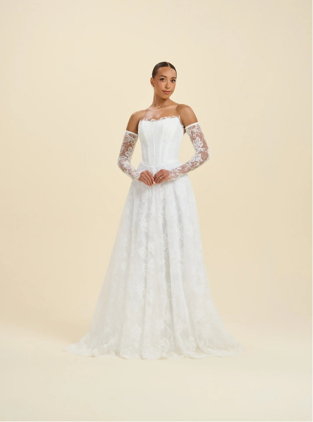 A woman modeling a white off-the-shoulder wedding gown with long sheer lace sleeves and a full lace-embroidered skirt, photographed against a plain warm cream studio background.
