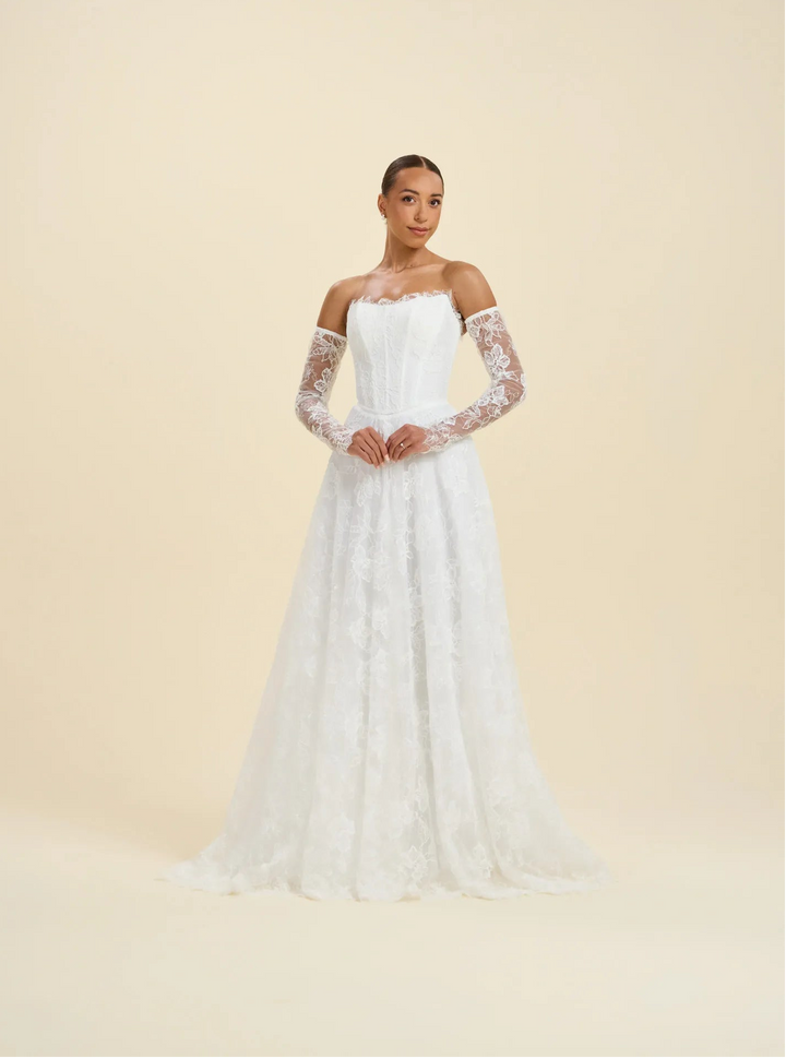 A woman modeling a white off-the-shoulder wedding gown with long sheer lace sleeves and a full lace-embroidered skirt, photographed against a plain warm cream studio background.