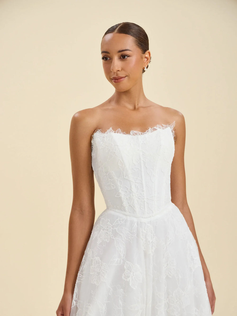A woman modeling a strapless white lace wedding dress against a warm beige studio background.