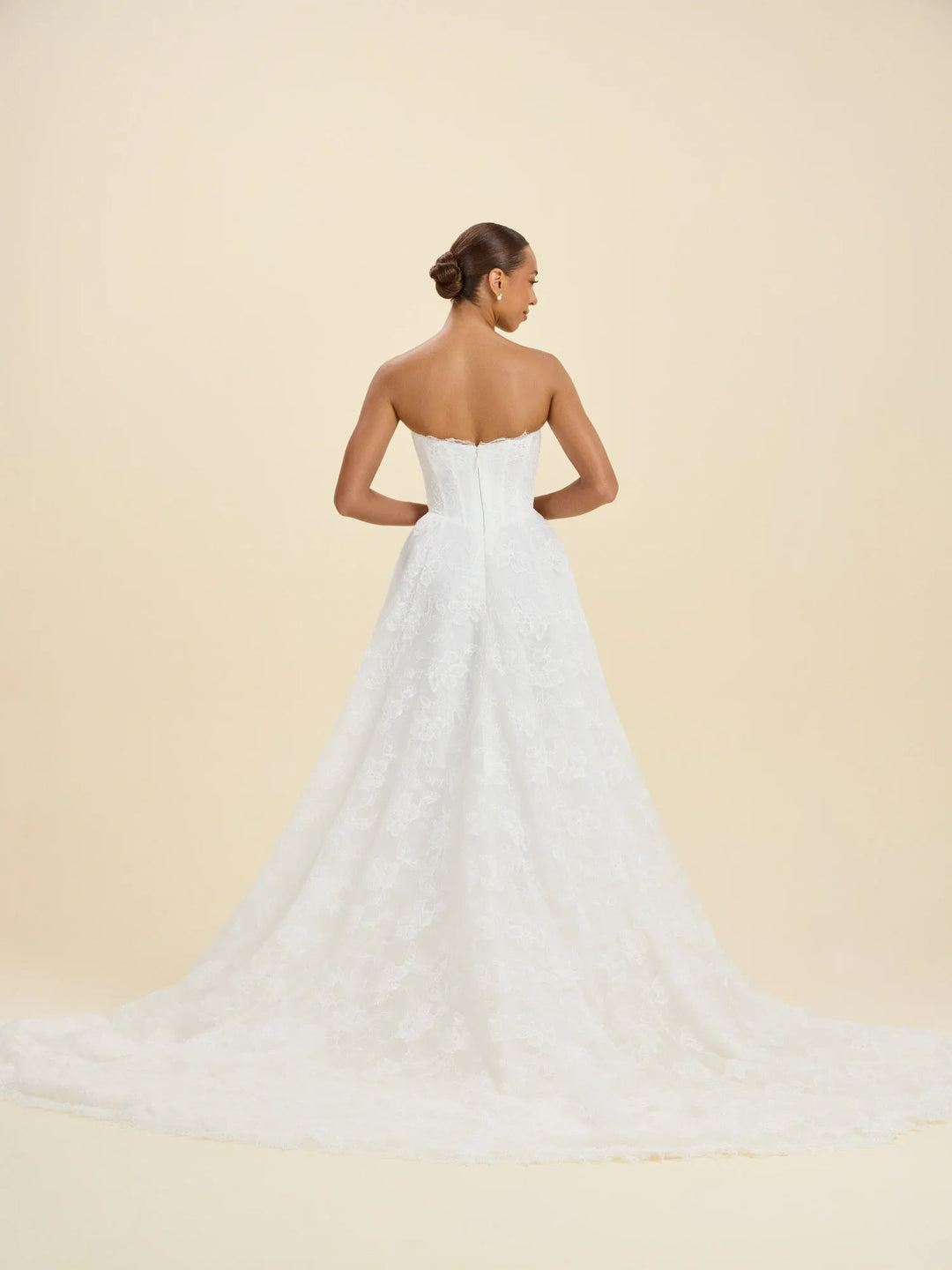 A woman modeling a strapless white lace wedding gown from the back, showing a structured backless bodice and a long flowing train, against a plain warm beige studio background.