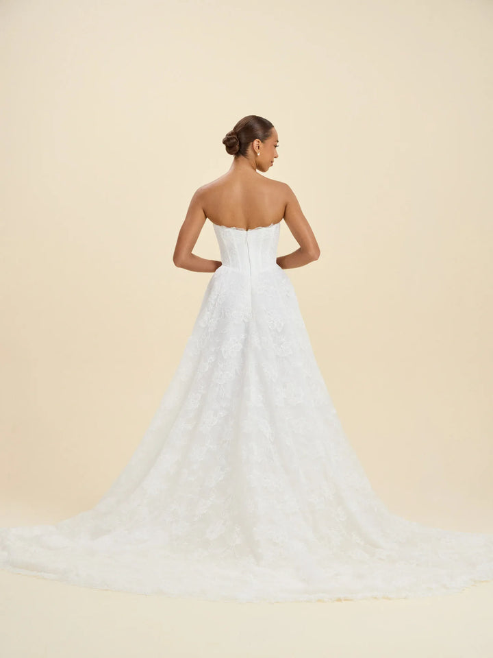 A woman modeling a strapless white lace wedding gown from the back, showing a structured backless bodice and a long flowing train, against a plain warm beige studio background.