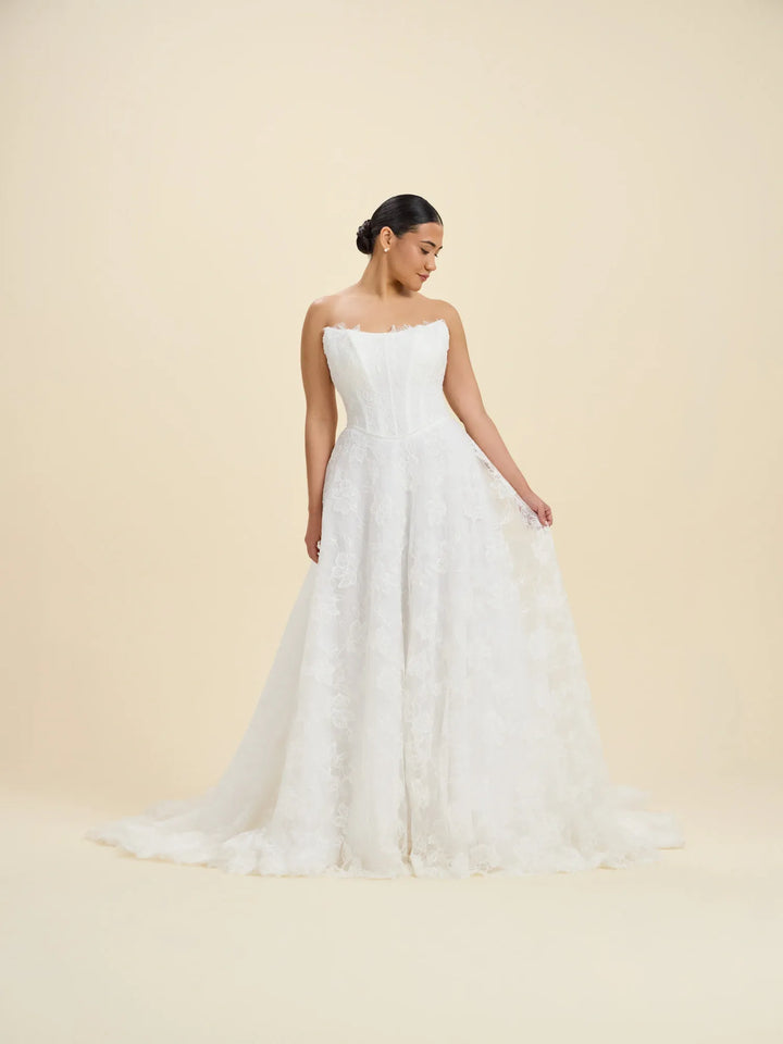A woman modeling a strapless white wedding gown with a fitted bodice and a full, textured tulle skirt against a plain beige studio background.