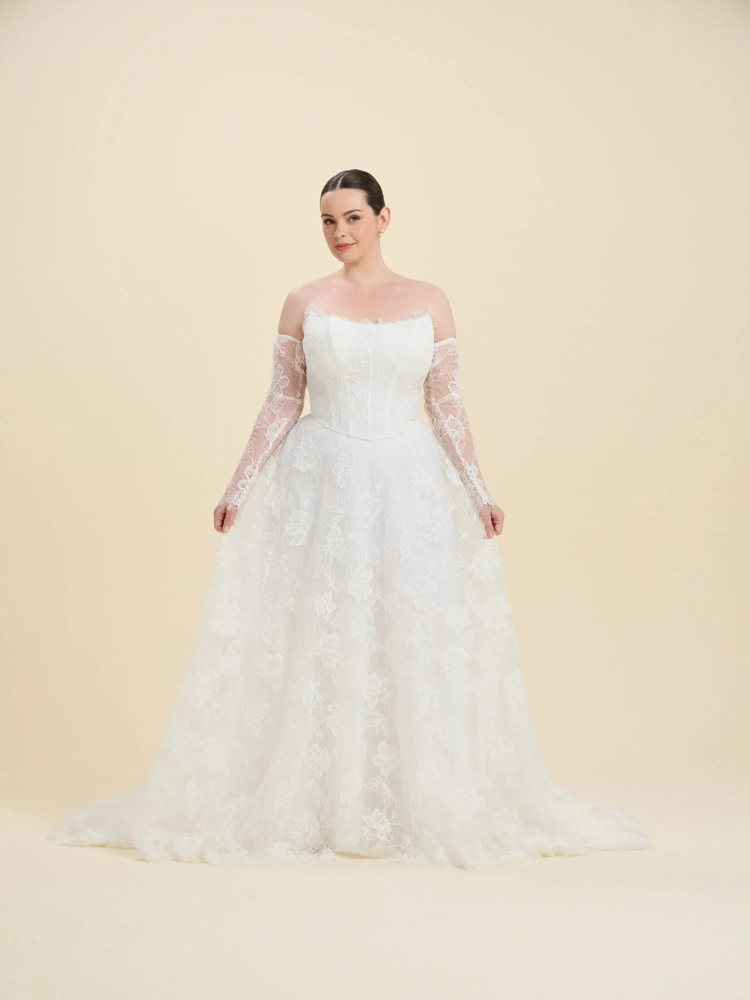 A woman modeling a white off-the-shoulder wedding gown with long sheer lace sleeves and a full lace-embroidered skirt, photographed against a plain warm beige studio background.