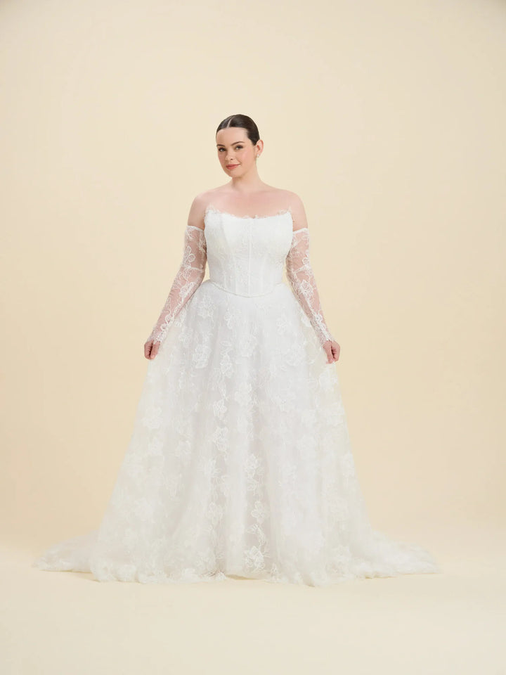 A woman modeling a white off-the-shoulder wedding gown with long sheer lace sleeves and a full lace-embroidered skirt, photographed against a plain warm beige studio background.