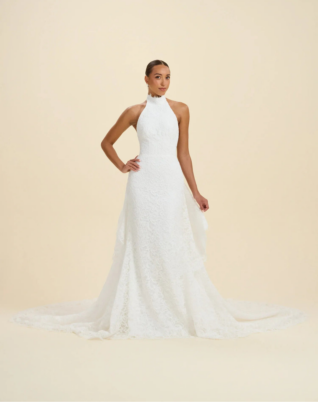 A woman modeling a sleeveless white wedding gown with a high halter neckline, fitted lace bodice, and a long train against a plain warm beige studio background.