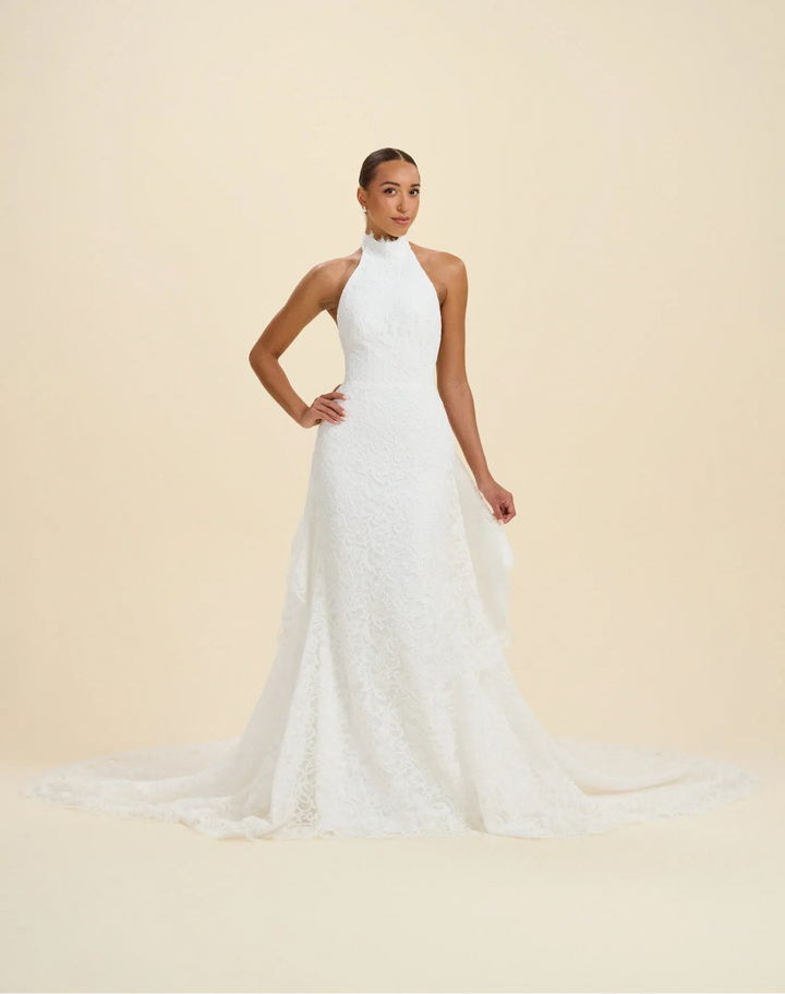 A woman modeling a sleeveless white wedding gown with a high halter neckline, fitted lace bodice, and a long train against a plain warm beige studio background.
