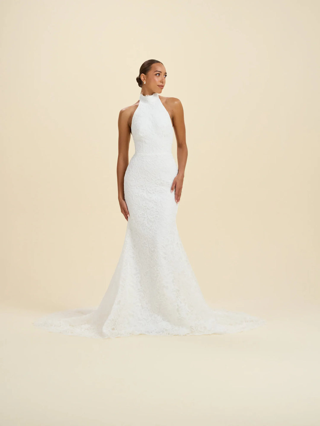 A woman modeling a sleeveless white lace wedding gown with a high halter-style neckline and a long train, photographed against a plain warm cream studio background.