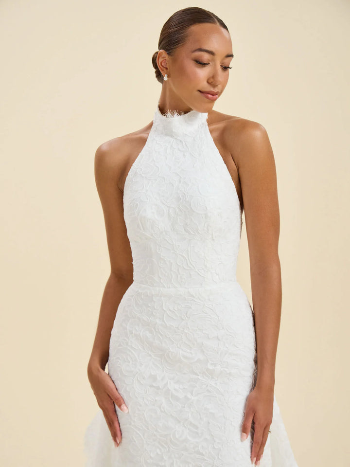 A woman modeling a white sleeveless halter-neck wedding dress with textured lace detailing, set against a solid beige background.