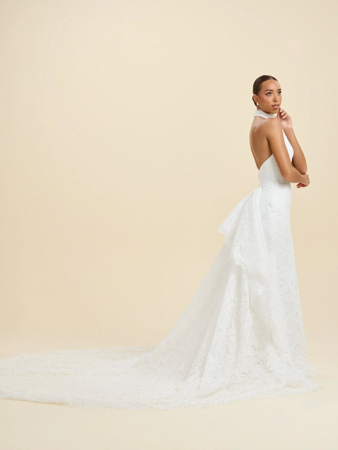 A woman modeling a white lace bridal gown from the back, featuring a high neck, fitted bodice, and a dramatic long train, set against a solid warm beige studio background.