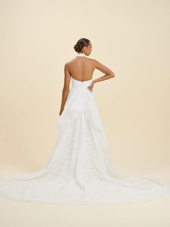 A woman wearing a white lace bridal gown is shown from the back against a warm beige studio background. The dress has a halter neckline, an open back, and a long train.